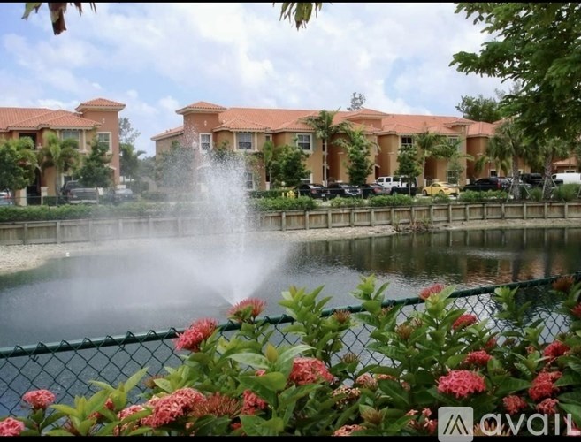 A fountain in the middle of a pond surrounded by flowers and houses in the background.