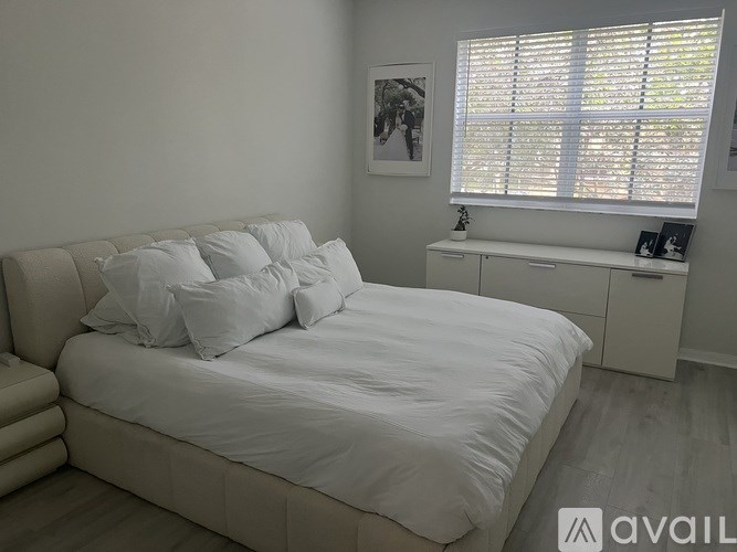 A white bed with a white headboard and white pillows in a room with a window covered in blinds.