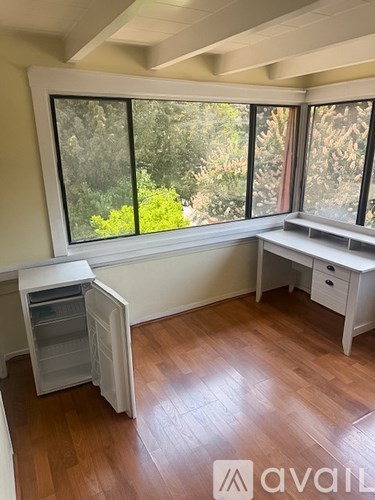 A room with a white fridge, a desk and a window with a view of trees.
