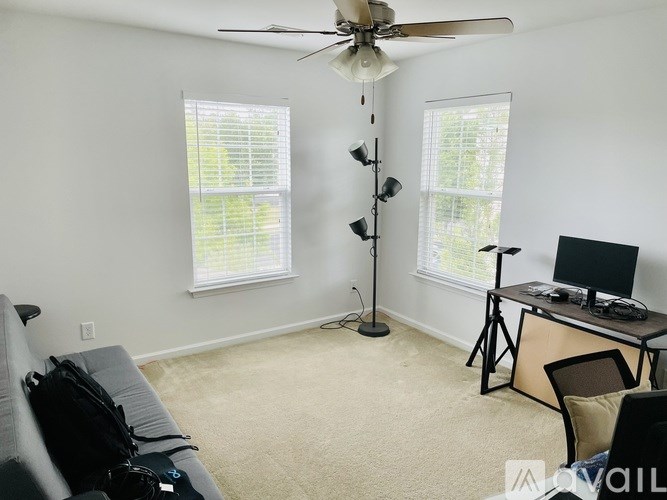 A room with a ceiling fan, two windows with blinds, a desk with a computer and a chair.