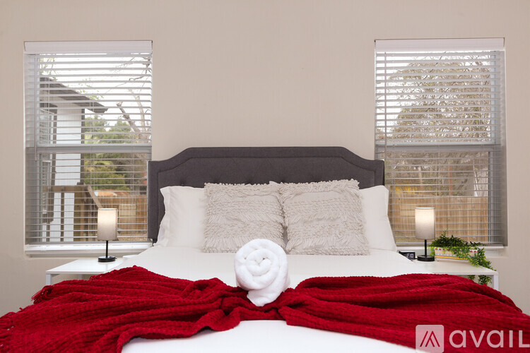 A bed with a red blanket and a white pillow is in the foreground with a window showing a view of a building in the background.
