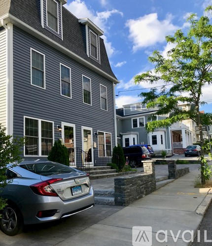 A silver car is parked on a sidewalk in front of a grey house.
