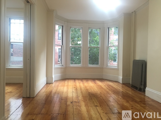 A room with wooden floors and white walls has a large window with a view of trees and a radiator on the wall.