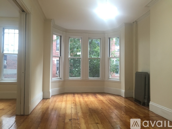 A room with wooden floors and white walls has a window with a view of trees.