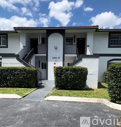 A white house with a black door and windows is surrounded by green bushes.