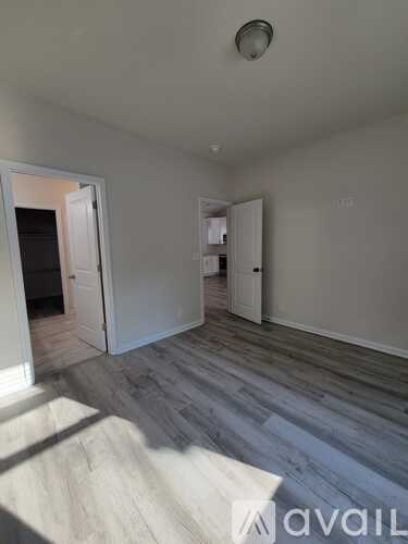 A room with wooden flooring and white walls, with a door open to another room.