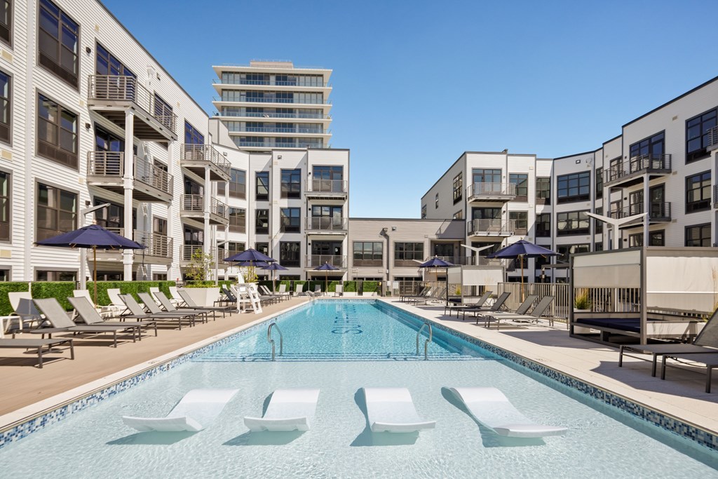 A swimming pool surrounded by sun loungers and umbrellas in front of apartment buildings.