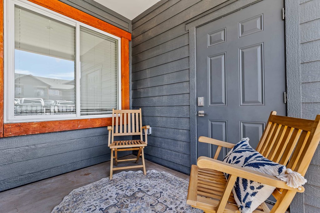 Two wooden chairs are on a porch with a grey door and a window.