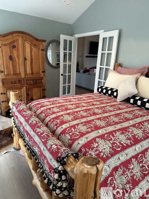 A bed with a red and white comforter is in the foreground of a room with a wooden armoire and a mirror.