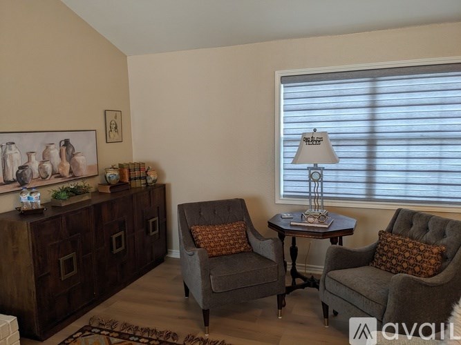 A living room with a grey sofa, a grey armchair, a wooden cabinet, a small table with a lamp, and a framed picture on the wall.