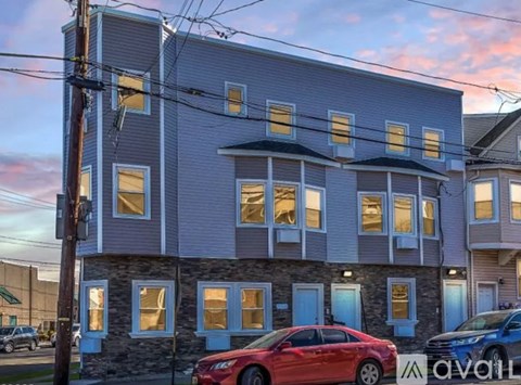 A red car is parked in front of a two-story building with a stone facade and multiple windows.