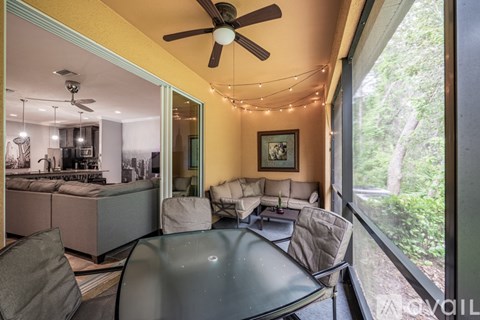 A living room with a glass table and chairs, a ceiling fan, and a view of the outdoors through a large window.