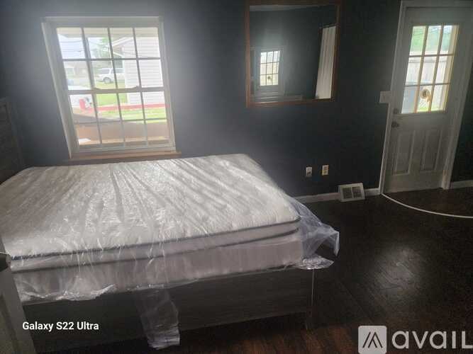 A bedroom with a bed covered in plastic and a window with a view of a house and trees outside.