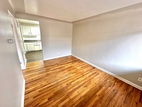 A room with wooden flooring and a kitchen visible through the door.