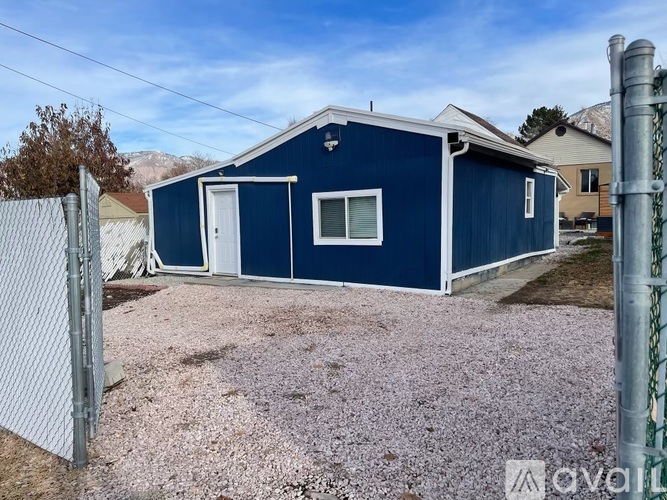 A blue house with a white door and windows is surrounded by a gravel driveway and a chain link fence.