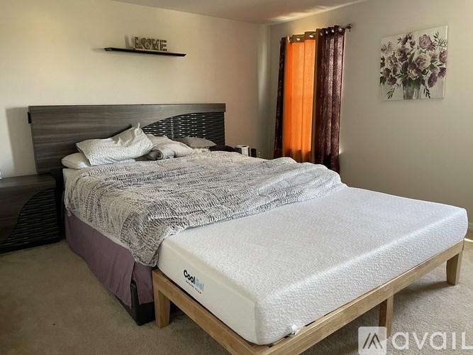 A bed with a wooden frame and a white mattress is in a room with a wall hanging and a window with orange curtains.
