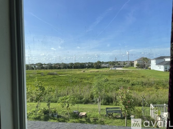 A view from a window looking out to a green field with a bench and a white house in the distance.