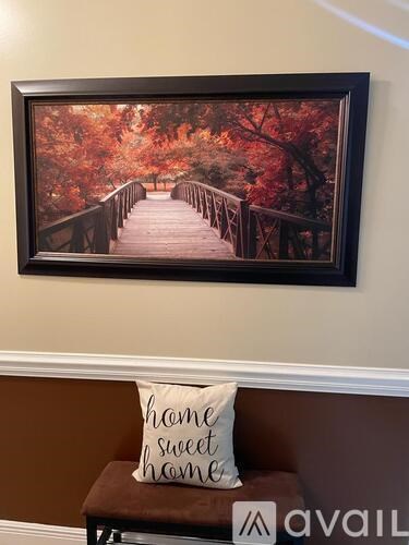 A framed picture of a bridge surrounded by trees in autumn is hanging on a wall above a bench with a pillow that says "home sweet home".