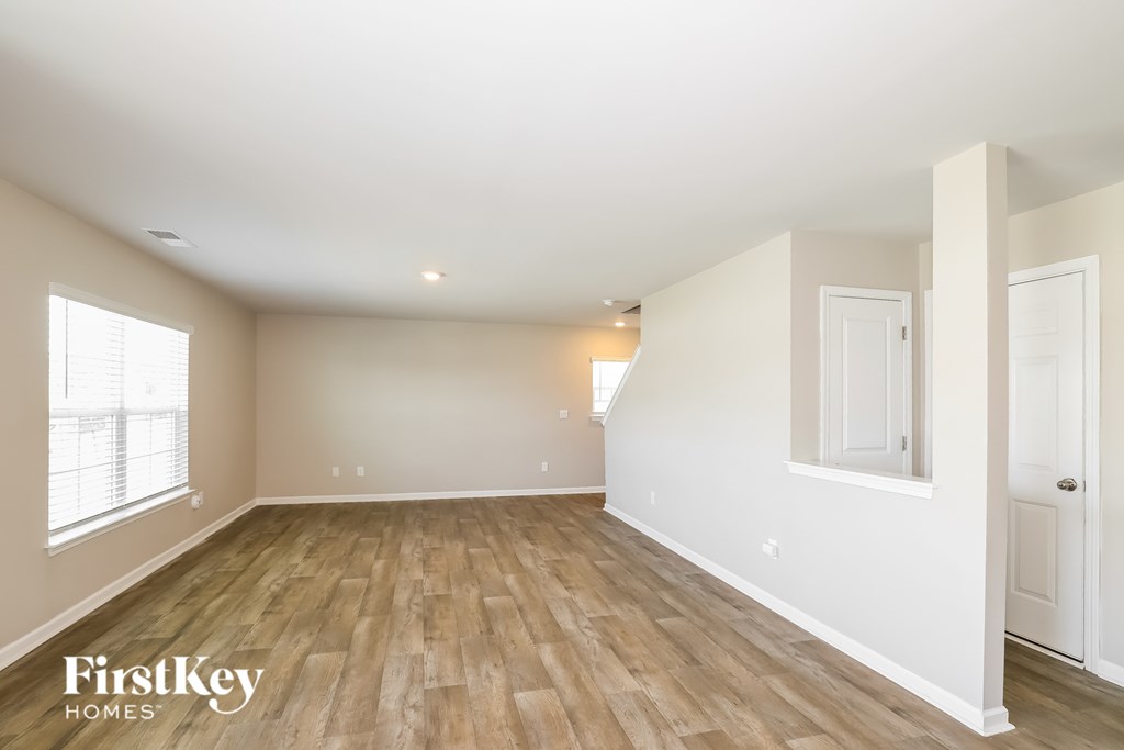 A spacious room with wooden flooring and white walls, with a window on the left side.