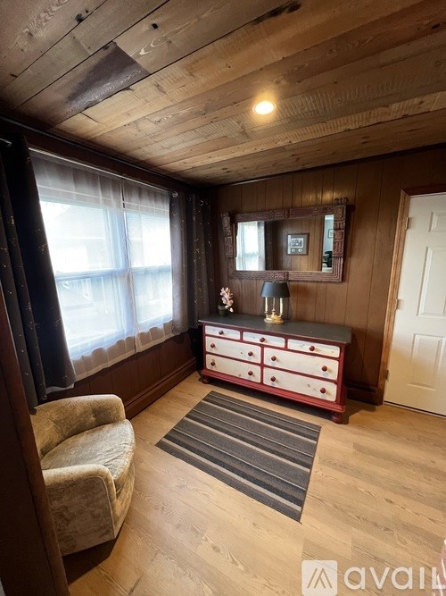 A room with wooden walls and ceiling, a white door, a grey rug, a wooden cabinet with a mirror and a vase on top, and a beige armchair.
