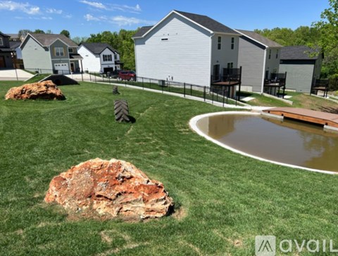 A large rock sits in the middle of a grassy area with a house and a pond in the background.