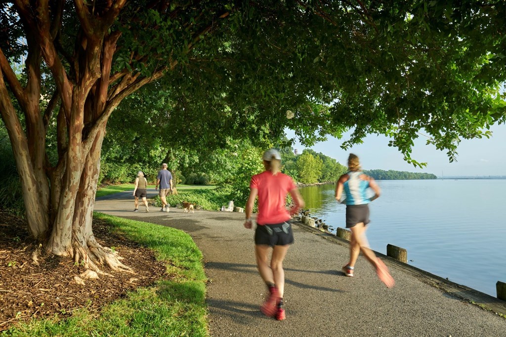 A woman in a red shirt and black shorts runs on a path next to a tree.
