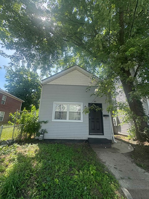 A small house with a grey siding and a black door is surrounded by greenery.