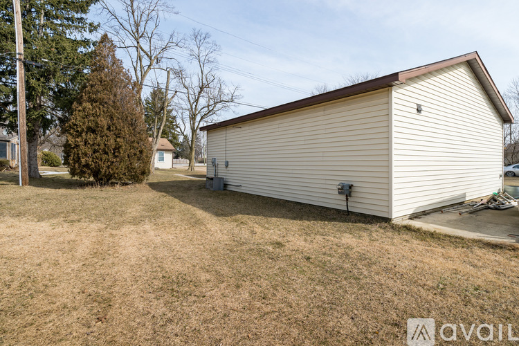 A large metal garage with a brown roof is situated in a grassy area with a tree and a house in the background.