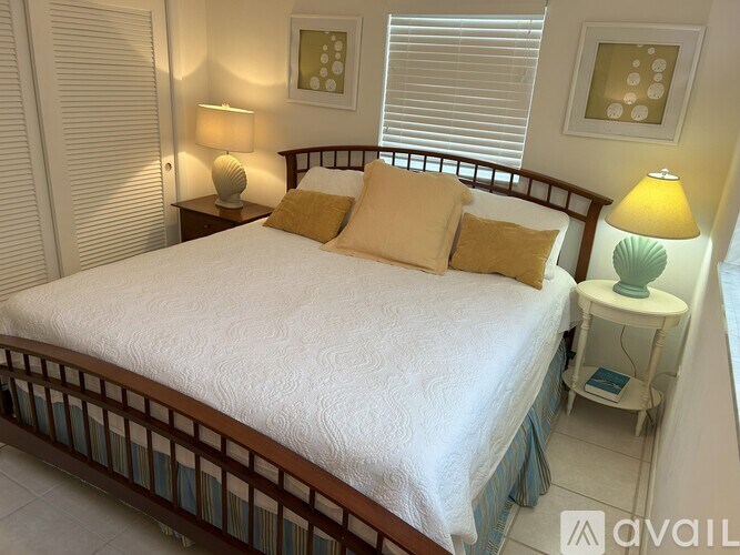 A bed with a white comforter and two yellow pillows sits in a room with a lamp and picture on the wall.
