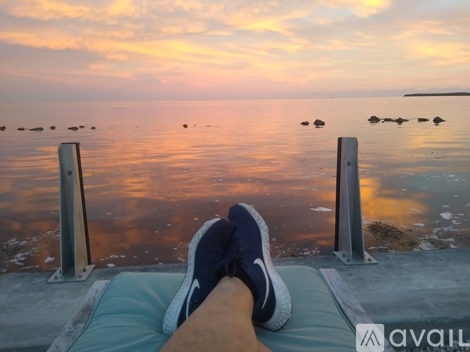 A person is sitting on a bench with their feet up, wearing blue sneakers, with a view of the sea and a sunset in the background.