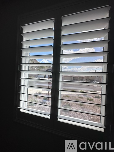 A window with white blinds showing a view of a cloudy sky and a building.