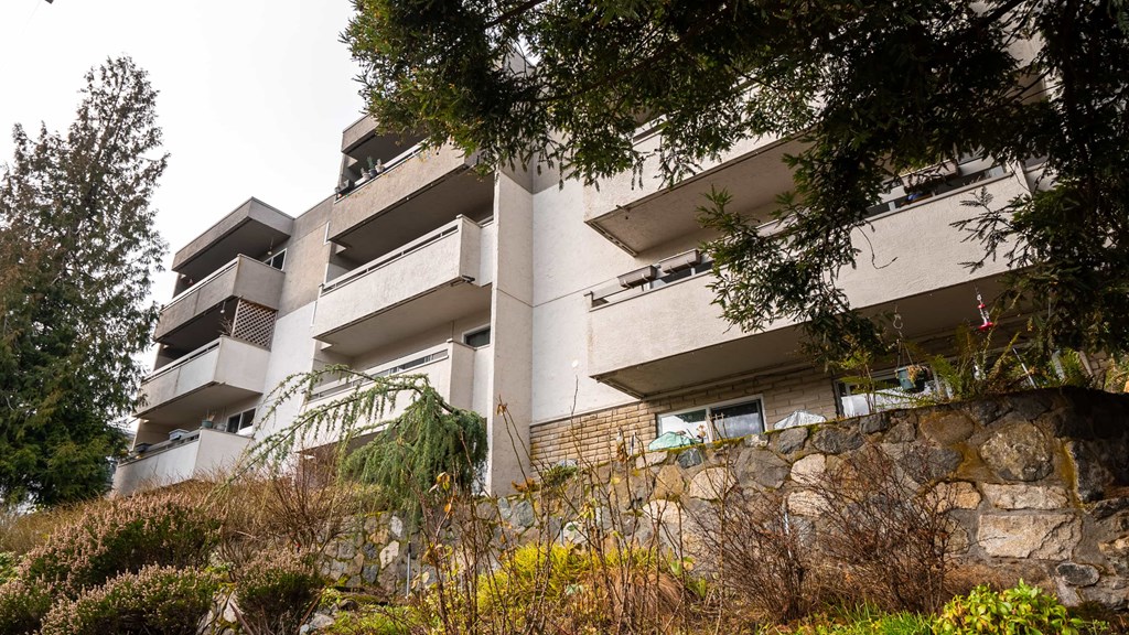 A large white building with balconies and windows is surrounded by trees and shrubs.