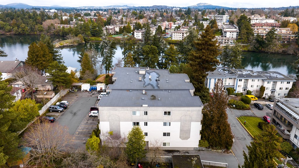 A white building with a grey roof is in the foreground of a cityscape with a lake and mountains in the background.