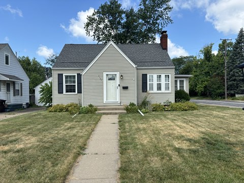 A small house with a grey roof and white door is surrounded by a grassy lawn.