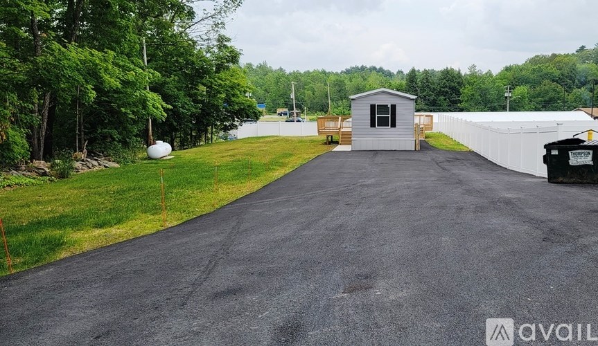 A gravel road leads to a small building with a white fence and trees in the background.