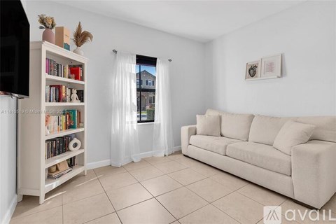 A living room with a cream sofa, a bookshelf filled with books, and a window with curtains.