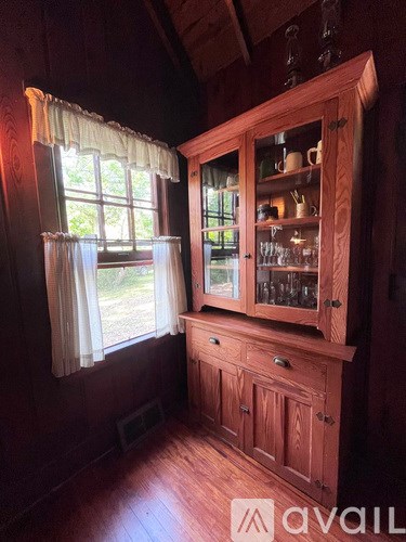 A wooden cabinet with glass doors and drawers is in a room with wooden flooring and a window with white curtains.