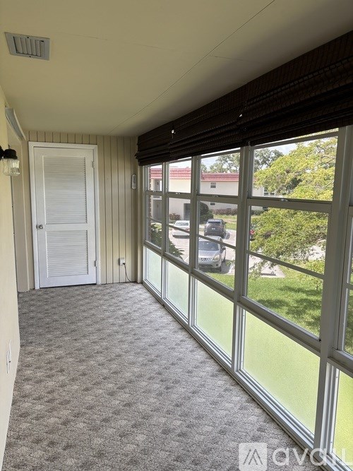 A room with a carpeted floor, a white door, and a large window with a view of a parking lot and trees.