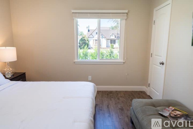 A bedroom with a bed, a nightstand with a lamp, a window with a view of the outside, and a door.