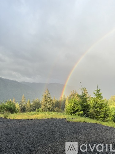 A rainbow appears over a road with trees and mountains in the background.