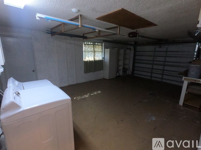 A white washing machine sits in a garage with a window covered in blinds.
