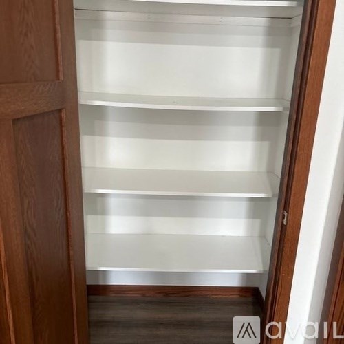 A white shelving unit with five shelves is in a room with a wooden floor and a wooden door.