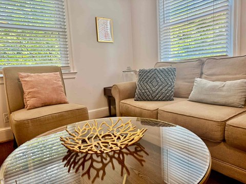 A living room with a beige couch, a chair, and a glass table with a wooden root-like object on it.