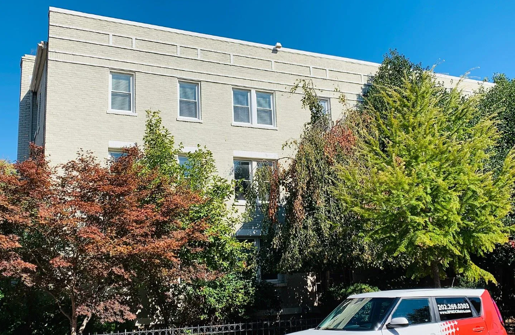 A red car is parked in front of a grey building with trees in the background.