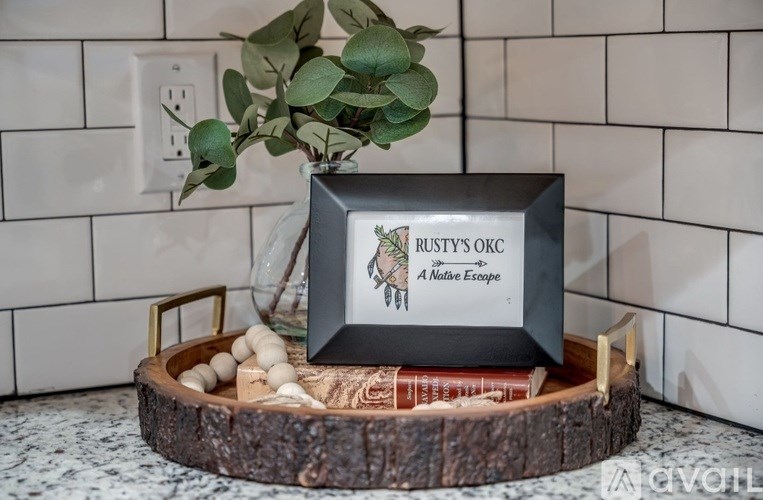 A picture frame with the text "RUSTY'S OKC A Nolene Escape" sits on a wooden tray with a plant and books.