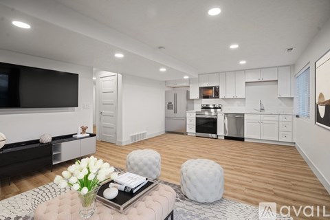 A modern living room with a flat screen TV, a coffee table with a vase of white flowers, and a kitchen in the background.