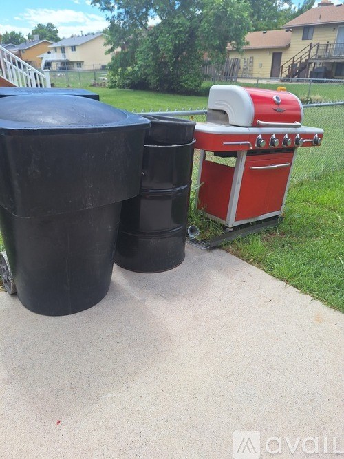 A red and white grill is on a concrete slab next to a black barrel.