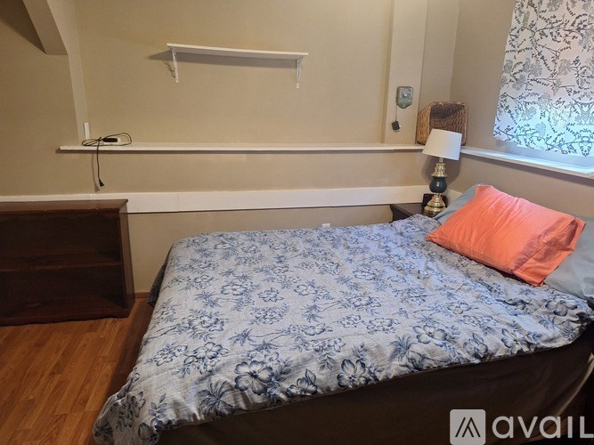 A bed with a blue floral comforter and an orange pillow sits in a room with a wooden dresser and a window with patterned curtains.