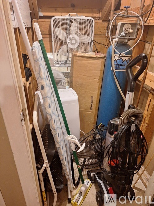 A cluttered room with a fan, a vacuum cleaner, and a painting on a stand.