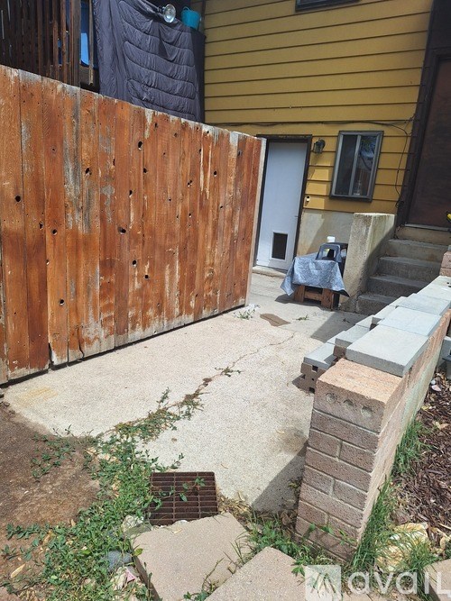 A wooden fence with a metal gate and a small concrete structure in front of a yellow house.